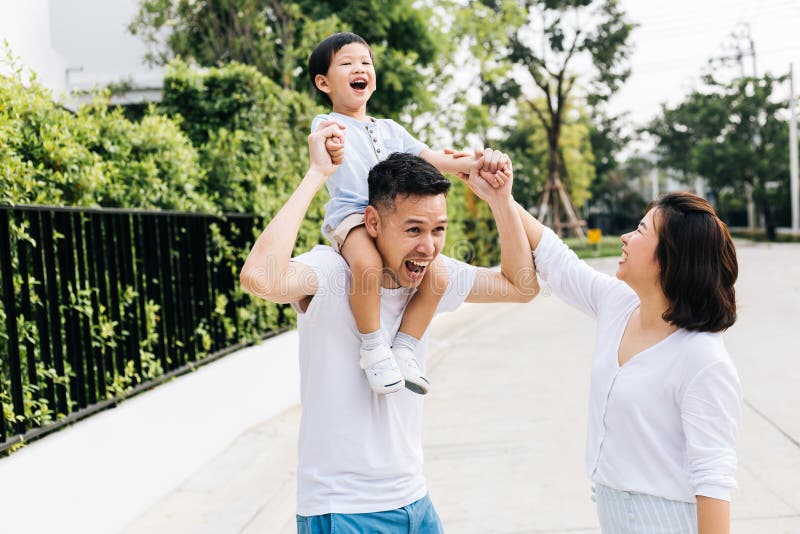 扛在肩上他的儿子的逗人喜爱的亚裔父亲与他的妻子一起在公园 举手的激动的家庭与幸福一起 免版税库存图片 扛在肩上他的儿子的逗人喜爱的亚裔父亲与他的妻子一起在公园 举手的激动的家庭与幸福一起 免版税库存图片