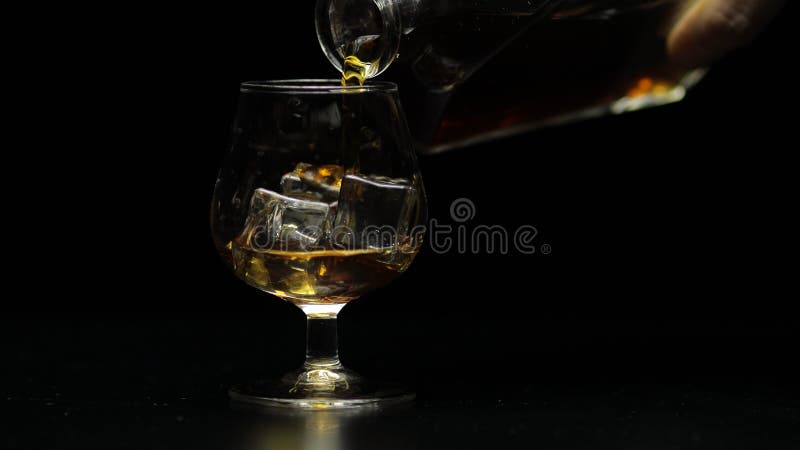 Pouring of golden whiskey, cognac or brandy from the bottle into glass with ice cubes. Black background. Side view of pouring glasses of alcohol drink bourbon, rum. Close-up shot. Pouring of golden whiskey, cognac or brandy from the bottle into glass with ice cubes. Black background. Side view of pouring glasses of alcohol drink bourbon, rum. Close-up shot