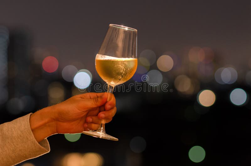 拿着一杯白酒的手敬酒对在屋顶酒吧的庆祝和党概念与城市光五颜六色的bokeh和 库存照片 拿着一杯白酒的手敬酒对在屋顶酒吧的庆祝和党概念与城市光五颜六色的bokeh和 库存照片