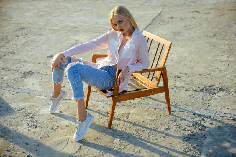 Sexy woman sit on wooden chair on concrete surface. Girl with sensual face makeup, blond hair on sunny day. Fashion, style concept. Beauty, look, youth. Summer vacation, leisure, lifestyle. Sexy woman sit on wooden chair on concrete surface. Girl with sensual face makeup, blond hair on sunny day. Fashion, style concept. Beauty, look, youth. Summer vacation, leisure, lifestyle.