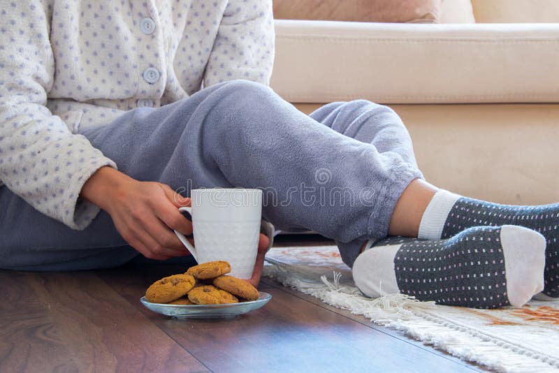 Cropped view of woman with pajamas sitting on the wooden floor at home with hot drink and cookies. Everyday cozy life concept. Cropped view of woman with pajamas sitting on the wooden floor at home with hot drink and cookies. Everyday cozy life concept