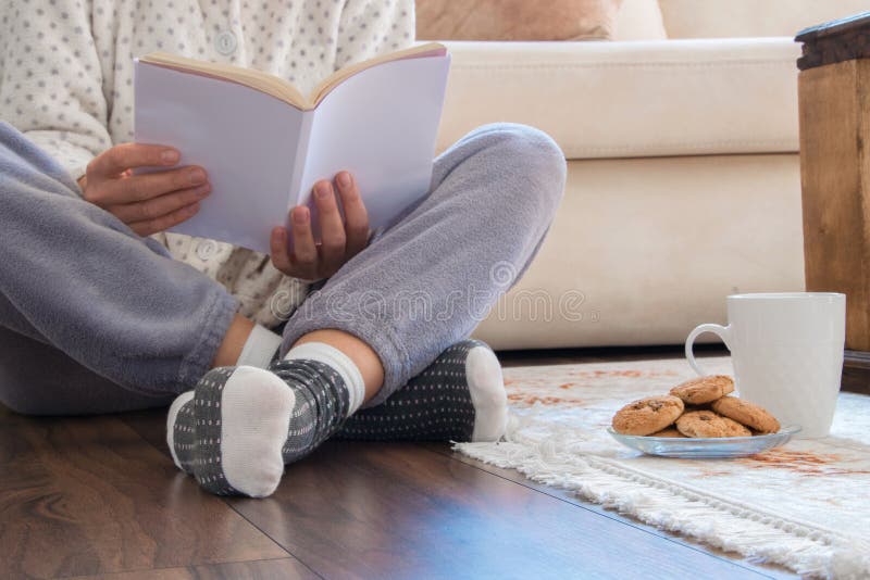 Cropped view of woman with pajamas sitting on the wooden floor at home with hot drink and cookies, reading a book- Everyday cozy life concept. Cropped view of woman with pajamas sitting on the wooden floor at home with hot drink and cookies, reading a book- Everyday cozy life concept