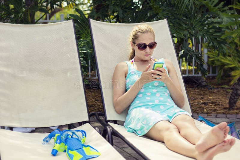 A beautiful woman sitting and relaxing in a lounge chair by the pool and reading her cell phone. A beautiful woman sitting and relaxing in a lounge chair by the pool and reading her cell phone