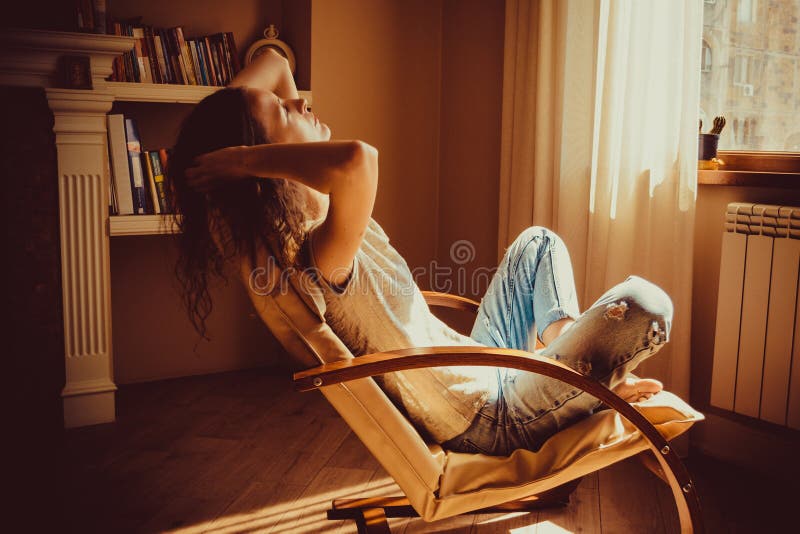 放松在舒适的现代椅子的工作以后的妇女在窗口附近在客厅 温暖的自然光 舒适家 便衣 C 库存照片