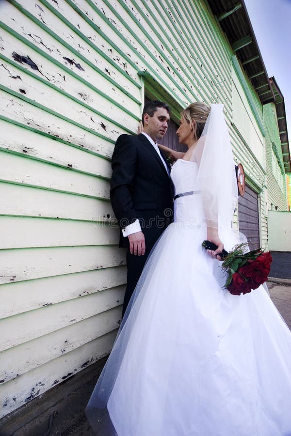 Outdoor portrait of a bride and groom by the side of a shingled building with peeling paint. Groom is standing against the building, while the bride is standing in front of him in an aggressive stance, with her right hand braced against the building over his shoulder. She is holding a bouquet of red roses in her left hand. Outdoor portrait of a bride and groom by the side of a shingled building with peeling paint. Groom is standing against the building, while the bride is standing in front of him in an aggressive stance, with her right hand braced against the building over his shoulder. She is holding a bouquet of red roses in her left hand.
