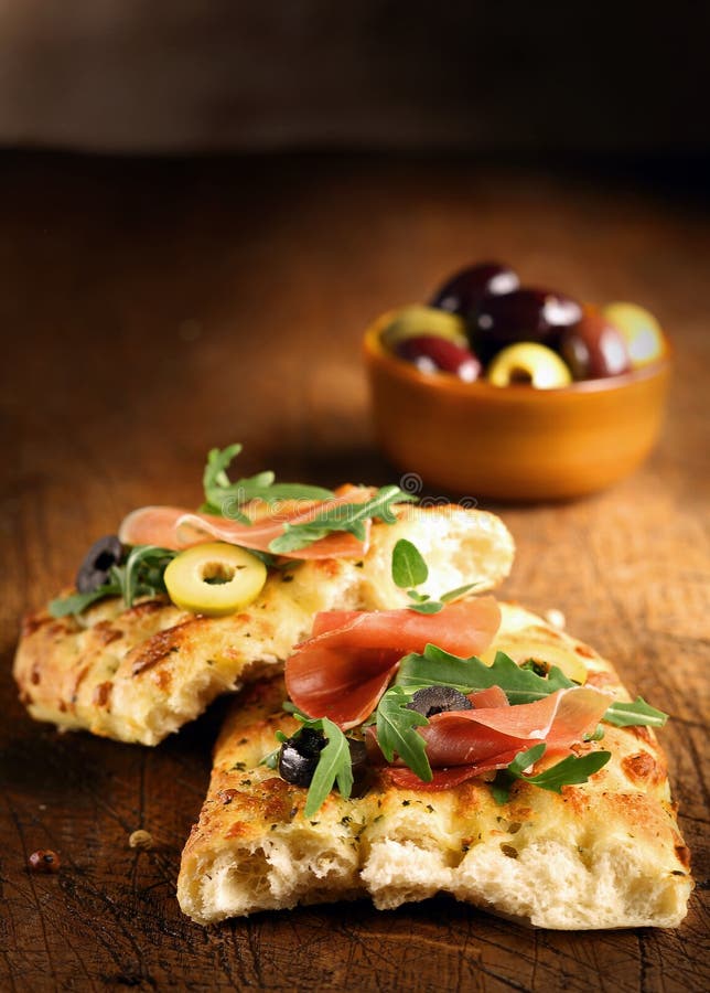 Serving of fresh traditional oven-baked Italian focaccia bread with ham, olives and rocket on a wooden table top with copyspace. Serving of fresh traditional oven-baked Italian focaccia bread with ham, olives and rocket on a wooden table top with copyspace