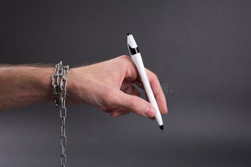 Unprofitable contracts concept, chained hand holding  pen ready to sign, close up, selective focus , blurred dark background. Unprofitable contracts concept, chained hand holding  pen ready to sign, close up, selective focus , blurred dark background