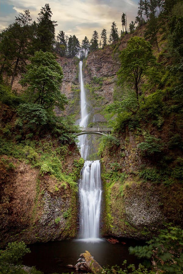 Multnomah falls, full view of both tiers, and bridge, early morning light and cloudy sky. Multnomah Falls is a waterfall located on Multnomah Creek in the Columbia River Gorge, east of Troutdale  Oregon, United States. The waterfall is accessible from the Historic Columbia River Highway and Interstate 84. Spanning two tiers on basalt cliffs, it is the tallest waterfall in the state of Oregon at 620 ft 189 m in height. Multnomah falls, full view of both tiers, and bridge, early morning light and cloudy sky. Multnomah Falls is a waterfall located on Multnomah Creek in the Columbia River Gorge, east of Troutdale  Oregon, United States. The waterfall is accessible from the Historic Columbia River Highway and Interstate 84. Spanning two tiers on basalt cliffs, it is the tallest waterfall in the state of Oregon at 620 ft 189 m in height.