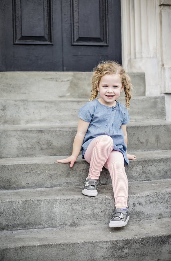 Emotional portrait of a happy little girl with blond curly hair. Smiling while sitting on the front steps of her home in an urban setting. Cute expression and adorable face. Carefree and Happiness concept photo. Emotional portrait of a happy little girl with blond curly hair. Smiling while sitting on the front steps of her home in an urban setting. Cute expression and adorable face. Carefree and Happiness concept photo