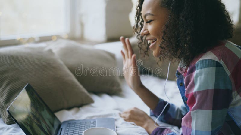 有卷曲非裔美国人的少妇与使用膝上型计算机照相机的朋友的录影闲谈，当说谎在床上时 免版税库存照片