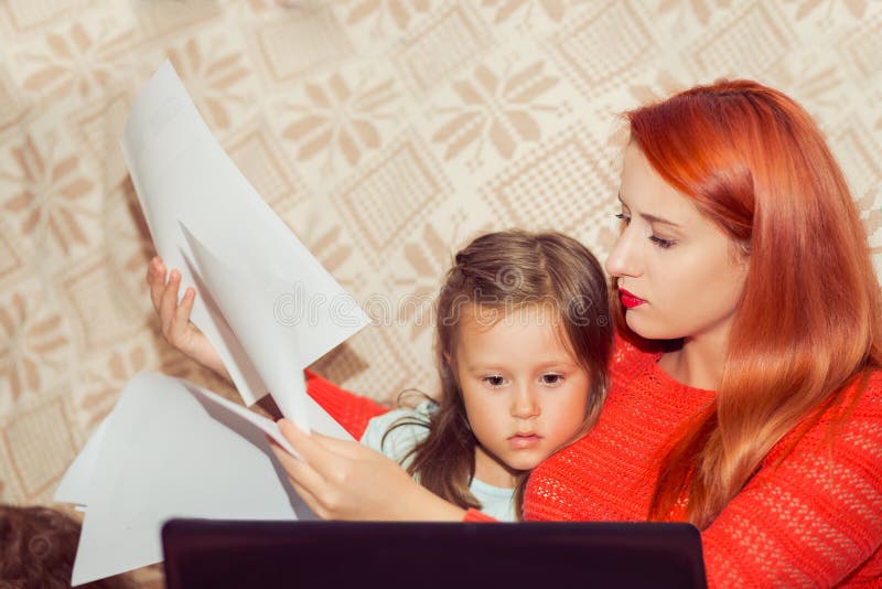 M with her child calculating home expenses. Busy M With child Running Business From Home looking at bank statement contract papers while kid looking cartoons on laptop sitting on bed. M with her child calculating home expenses. Busy M With child Running Business From Home looking at bank statement contract papers while kid looking cartoons on laptop sitting on bed