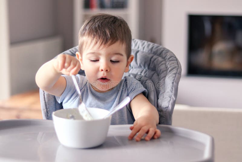 有滑稽的被抹上的面孔的逗人喜爱的矮小的男婴集中食物吃与从白色碗的叉子在他前面的桌上sittin的 库存照片 有滑稽的被抹上的面孔的逗人喜爱的矮小的男婴集中食物吃与从白色碗的叉子在他前面的桌上sittin的 库存照片