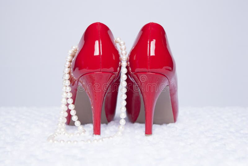 A pair of sexy, red patent leather pumps sit atop a white fur surface with a white background; pearls are draped over the side of one shoe. A pair of sexy, red patent leather pumps sit atop a white fur surface with a white background; pearls are draped over the side of one shoe