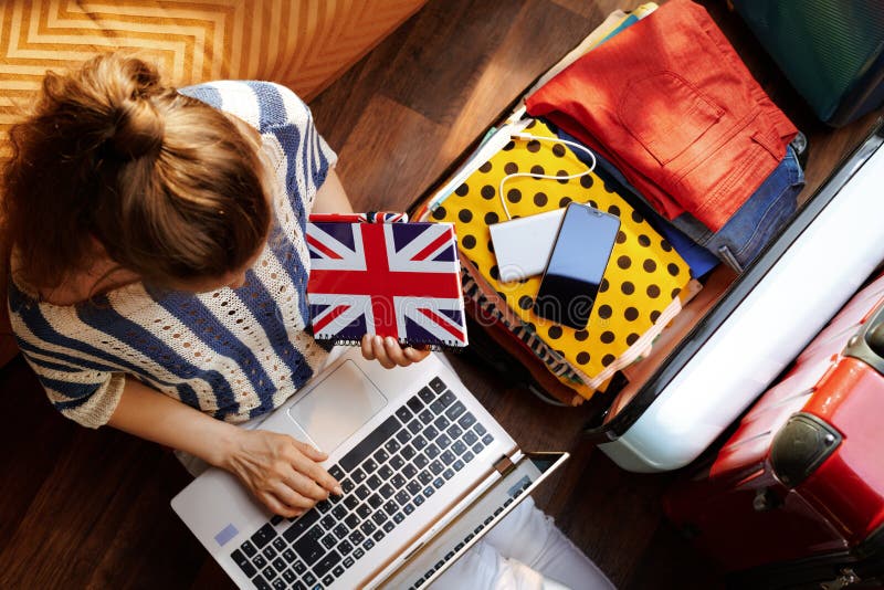 æœ‰è‹±å›½å›½æ——ç¬”è®°æœ¬çš„å¥³äººä¹°æœºç¥¨ã€‚é˜³å…‰æ˜Žåªšçš„å¤æ—¥ï¼ŒçŽ°ä»£å®¢åŽ…é‡Œï¼Œæ—¶å°šå¥³æ€§ç©¿ç™½è‰²è£¤å­ã€æ¡çº¹è¡¬è¡«ã 库存照片