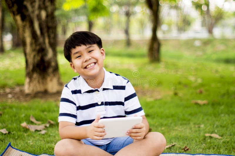 有逗人喜爱的微笑的亚裔男孩,与在手中片剂,站立在庭院、网上学习的概念和电子教学里与学会外部 库存照片 有逗人喜爱的微笑的亚裔男孩,与在手中片剂,站立在庭院、网上学习的概念和电子教学里与学会外部 库存照片