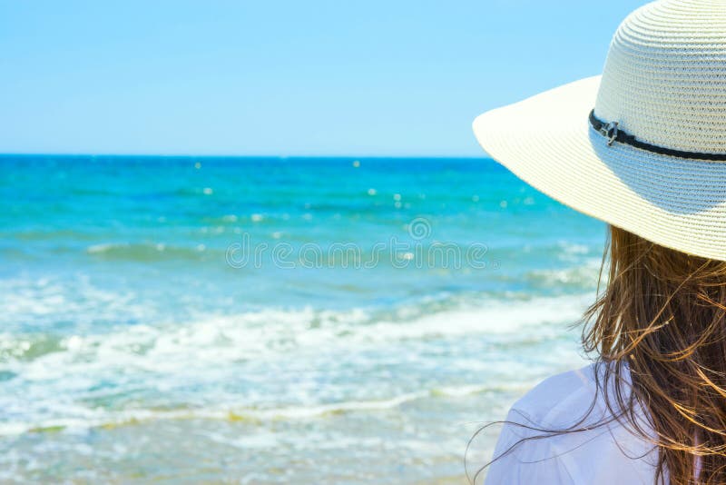 有长的栗子头发的年轻可爱的白种人妇女在与后面的帽子立场在沙子海滩看绿松石海天线 免版税图库摄影 有长的栗子头发的年轻可爱的白种人妇女在与后面的帽子立场在沙子海滩看绿松石海天线 免版税图库摄影