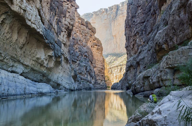 The Rio Grande River running through Santa Elena Canyon at Big Bend National Park in Texas. A hiker rests by the riverbank in the lower right, facing away from the camera. The Rio Grande River running through Santa Elena Canyon at Big Bend National Park in Texas. A hiker rests by the riverbank in the lower right, facing away from the camera.