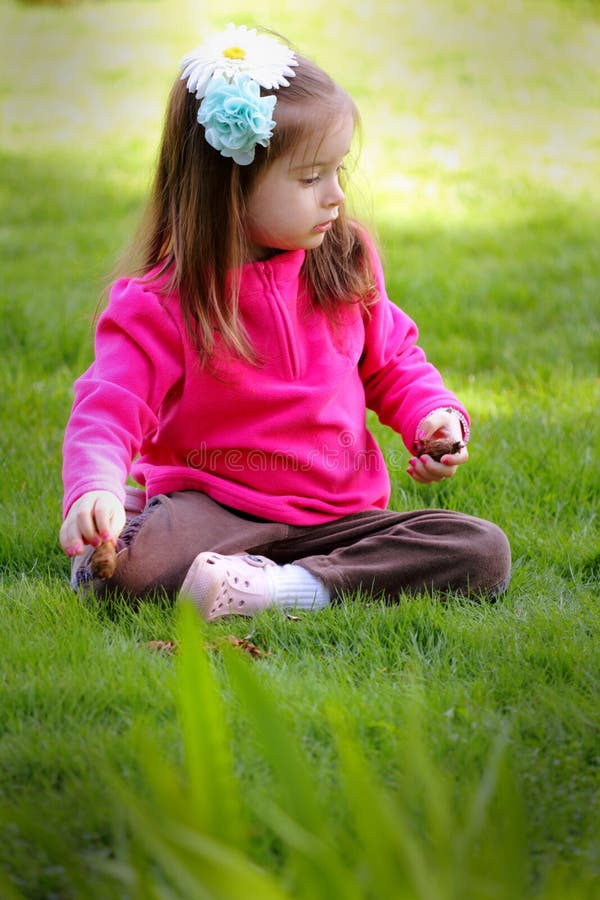 A cute happy little girl in pink with painted fingernails and bows in her long dark hair sitting in the yard playing. Shallow depth of field. A cute happy little girl in pink with painted fingernails and bows in her long dark hair sitting in the yard playing. Shallow depth of field.