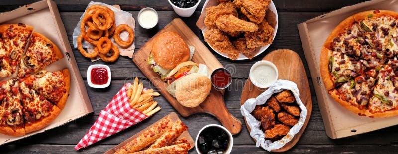 Table scene of assorted take out or delivery foods. Hamburgers, pizza, fried chicken and sides. Top down view on a dark wood banner background. Table scene of assorted take out or delivery foods. Hamburgers, pizza, fried chicken and sides. Top down view on a dark wood banner background