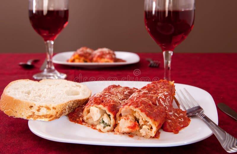Two plates of stuffed manicotti, a loaf of sliced Italian bread and 2 glasses of wine. Two plates of stuffed manicotti, a loaf of sliced Italian bread and 2 glasses of wine