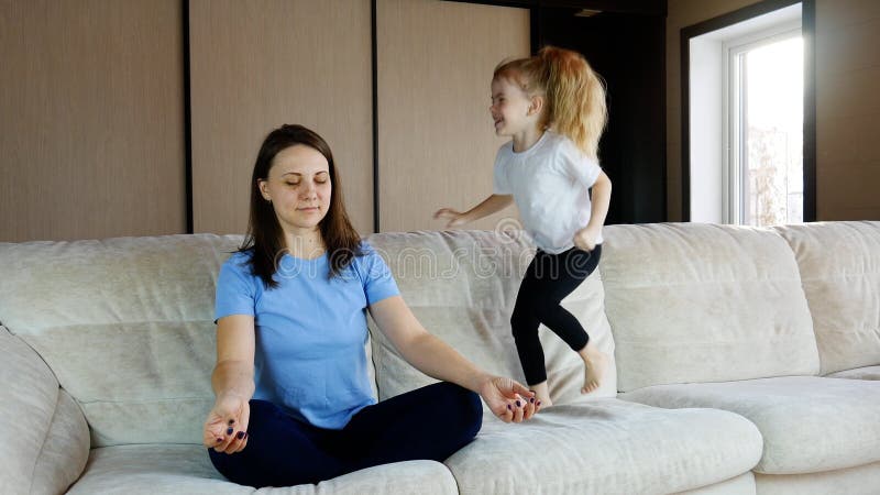 M meditating sitting on sofa while active energetic child daughter jumping playing, calm young mom doing yoga exercise at home for stress relief relaxing with naughty little kid. M meditating sitting on sofa while active energetic child daughter jumping playing, calm young mom doing yoga exercise at home for stress relief relaxing with naughty little kid