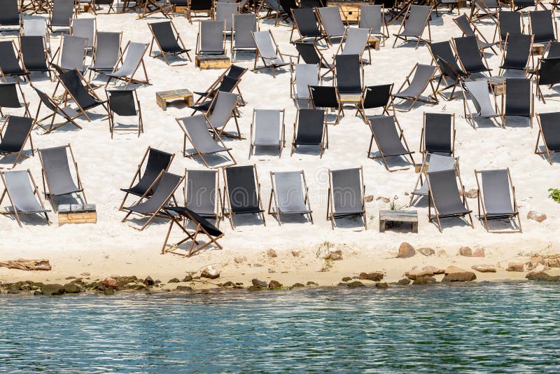 Scenic water view of many empty black wooden deckchair loungers on sand river or sea ocean beach. Rows of deckchair on closed coast due contamination. Lack of people tourist on resort due sanctions. Scenic water view of many empty black wooden deckchair loungers on sand river or sea ocean beach. Rows of deckchair on closed coast due contamination. Lack of people tourist on resort due sanctions.