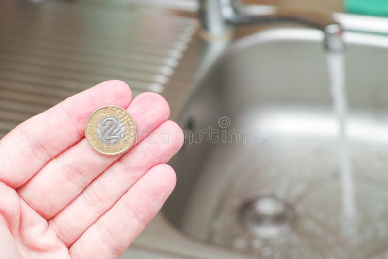 Hand holding a two zloty coin infront of a running faucet - for concepts like cheap water, savings potential at home, household spendings or wasting water - focus is on the 2 zloty coin. Hand holding a two zloty coin infront of a running faucet - for concepts like cheap water, savings potential at home, household spendings or wasting water - focus is on the 2 zloty coin