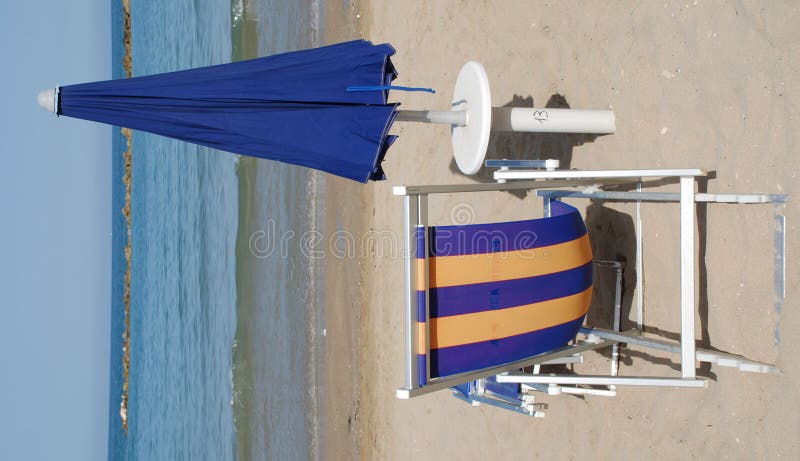 Closed blue beach umbrellas on a beach on a sunny summers day next to a deckchair. Closed blue beach umbrellas on a beach on a sunny summers day next to a deckchair
