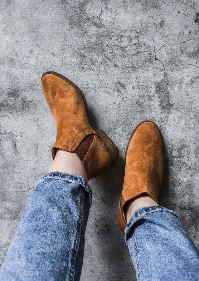 Women`s feet in mom`s jeans and chelsea suede boots on a gray background, top view. Women`s feet in mom`s jeans and chelsea suede boots on a gray background, top view.