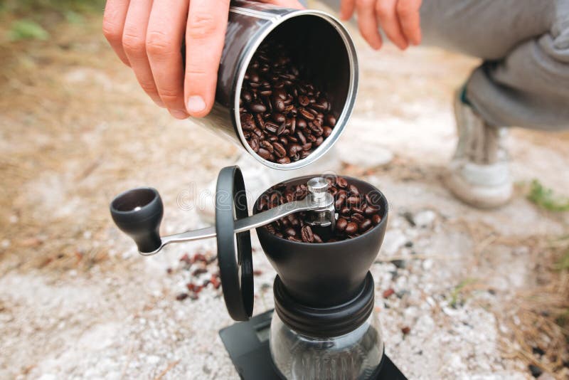 Filling grinder with fresh coffee beans on cliff at lake, preparing for brewing alternative coffee at camping. Making hot drink at picnic outdoors. Trekking and hiking in mountains. Filling grinder with fresh coffee beans on cliff at lake, preparing for brewing alternative coffee at camping. Making hot drink at picnic outdoors. Trekking and hiking in mountains