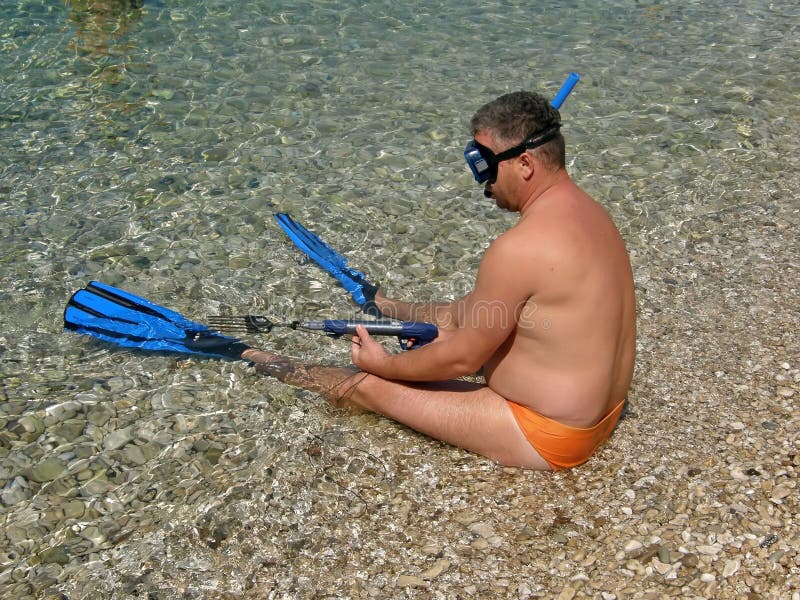 Alone diver with harpoon sitting on pebble beach in a shallow at Adriatic sea at sunny day. Horizontal color photo. Alone diver with harpoon sitting on pebble beach in a shallow at Adriatic sea at sunny day. Horizontal color photo.