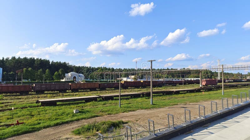 Railway tracks have been laid and electrified at the station with administrative buildings among the grassy lawns. There are trains of freight wagons and platforms on them. There is a forest behind the station. Sunny. Railway tracks have been laid and electrified at the station with administrative buildings among the grassy lawns. There are trains of freight wagons and platforms on them. There is a forest behind the station. Sunny