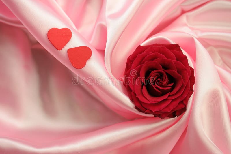 Valentine's Day theme - a detail of a red rose and two little red hearts lying on pink satin fabric. Valentine's Day theme - a detail of a red rose and two little red hearts lying on pink satin fabric.