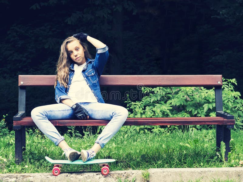 牛仔裤衬衣的美丽的白肤金发的青少年的女孩,坐与背包和滑板的长凳在公园 库存图片 牛仔裤衬衣的美丽的白肤金发的青少年的女孩,坐与背包和滑板的长凳在公园 库存图片