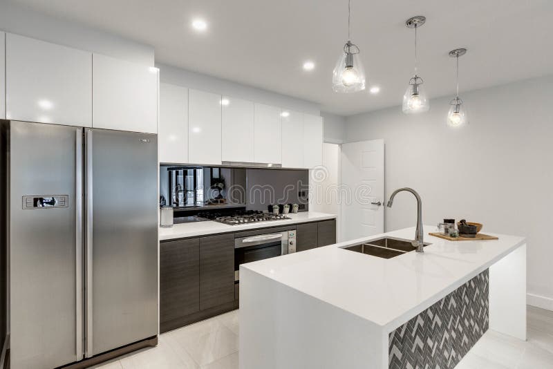 CANBERRA, AUSTRALIA â€“ JUNE 2, 2018: Beautiful new kitchen furnished with stainless steel fridge, reflective splashback, island, inset sink, stove, oven and three lit hanging lights in a modern home. CANBERRA, AUSTRALIA â€“ JUNE 2, 2018: Beautiful new kitchen furnished with stainless steel fridge, reflective splashback, island, inset sink, stove, oven and three lit hanging lights in a modern home