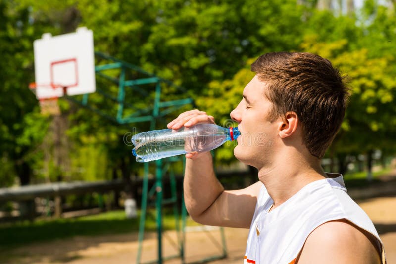 Head and Shoulders View of Young Man Drinking Water from Bottle, Taking a Break for Refreshment and Hydration on Basketball Court. Head and Shoulders View of Young Man Drinking Water from Bottle, Taking a Break for Refreshment and Hydration on Basketball Court