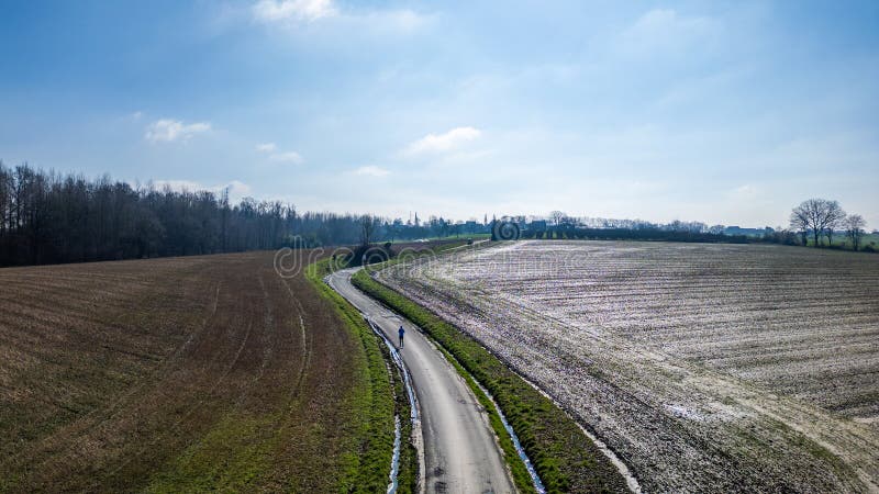 The image presents an aerial view of a curvy, narrow road meandering through expansive fields. The fields appear dormant with remnants of snow or frost, suggesting this photo was taken during colder months. A line of trees can be seen in the distance, marking the horizon under a mostly clear blue sky with minimal clouds. The road serves as a leading line, drawing the eye through the composition, and there are no visible vehicles or people, giving a sense of solitude and tranquility. Aerial Perspective of Curved Road Amidst Fields. High quality photo. The image presents an aerial view of a curvy, narrow road meandering through expansive fields. The fields appear dormant with remnants of snow or frost, suggesting this photo was taken during colder months. A line of trees can be seen in the distance, marking the horizon under a mostly clear blue sky with minimal clouds. The road serves as a leading line, drawing the eye through the composition, and there are no visible vehicles or people, giving a sense of solitude and tranquility. Aerial Perspective of Curved Road Amidst Fields. High quality photo