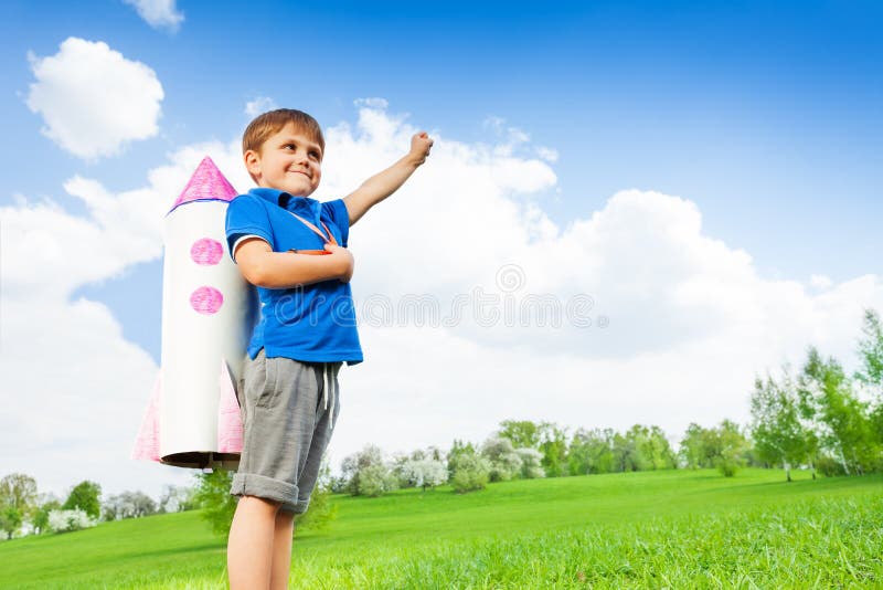 Boy wears paper rocket toy on his shoulders and holds arm straight up during sunny day in the green field. Boy wears paper rocket toy on his shoulders and holds arm straight up during sunny day in the green field