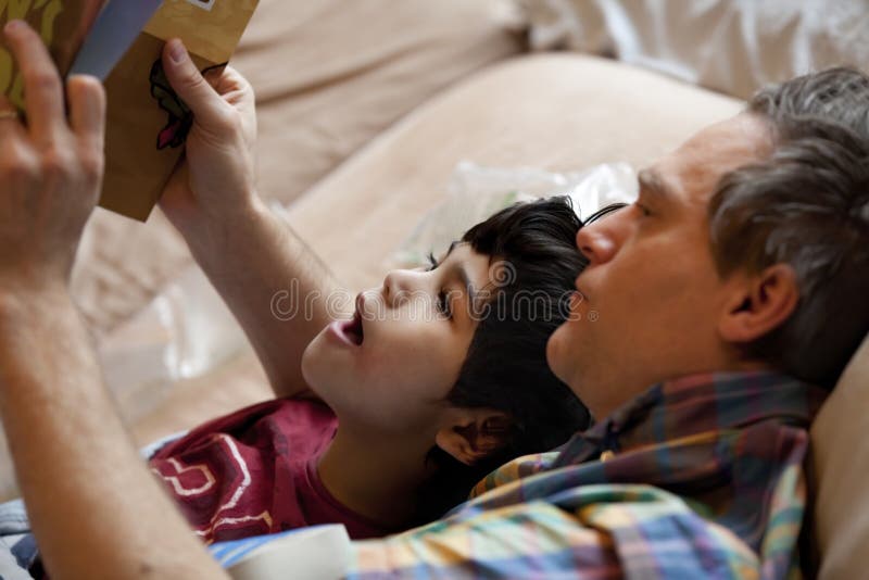 男孩躺在床上与父亲一起读书 库存图片