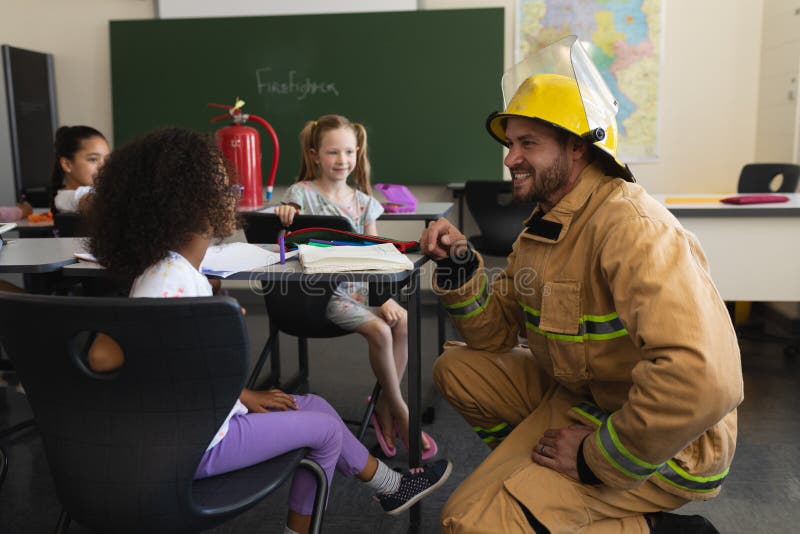 Side view of male Caucasian firefighter on his knees teaching schoolgirls about fire safety with smile in classroom of elementary school. Side view of male Caucasian firefighter on his knees teaching schoolgirls about fire safety with smile in classroom of elementary school