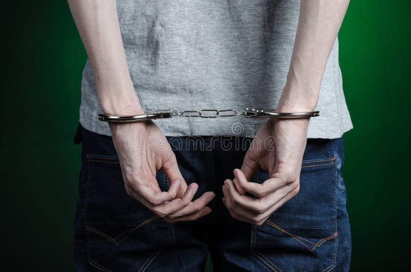 Prison and convicted topic: man with handcuffs on his hands in a gray T-shirt and blue jeans on a dark green background, put handcuffs on the drug dealer, the view from the back. Prison and convicted topic: man with handcuffs on his hands in a gray T-shirt and blue jeans on a dark green background, put handcuffs on the drug dealer, the view from the back