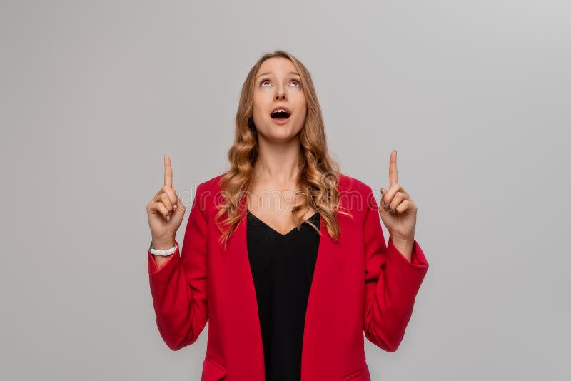 Hey look there. Excited blonde young woman pointing fingers up, looking amazed, showing something interesting, stands in red blazer against gray background. Hey look there. Excited blonde young woman pointing fingers up, looking amazed, showing something interesting, stands in red blazer against gray background