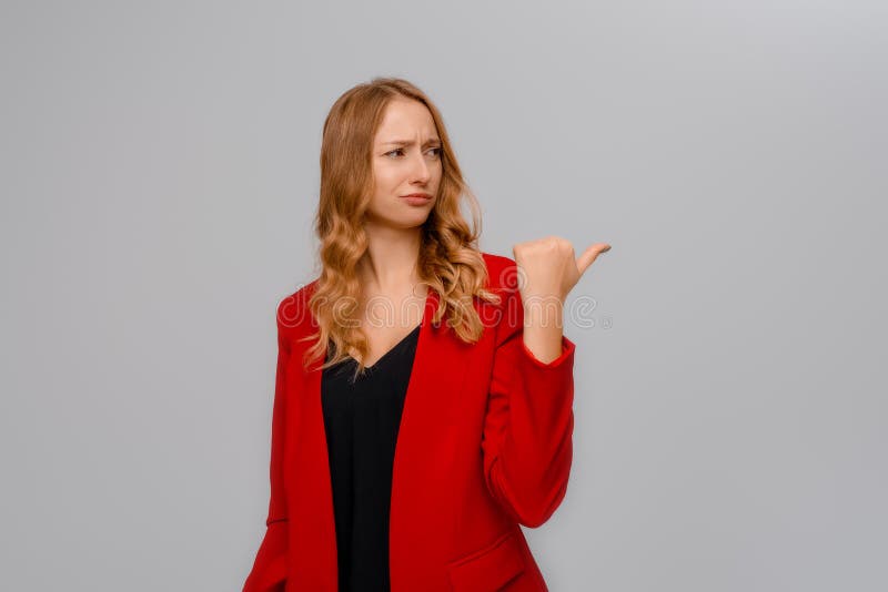 Hey look there. Disappointed blonde young woman pointing finger to the right, looking upset and frowning skeptical, showing bad offer, standing in red blazer against gray background. Hey look there. Disappointed blonde young woman pointing finger to the right, looking upset and frowning skeptical, showing bad offer, standing in red blazer against gray background