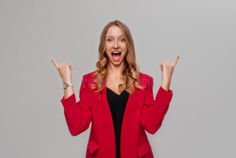 Hey look there. Smiling excited blonde young woman pointing fingers up, showing to advertisement, promo offer, stands in red blazer against gray background. Hey look there. Smiling excited blonde young woman pointing fingers up, showing to advertisement, promo offer, stands in red blazer against gray background