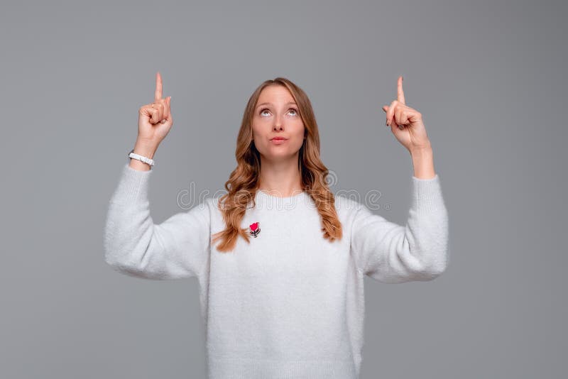 Hey look there. Beautiful blonde young woman pointing fingers up, looking amazed, showing something interesting, stands in white sweater against gray background. Hey look there. Beautiful blonde young woman pointing fingers up, looking amazed, showing something interesting, stands in white sweater against gray background