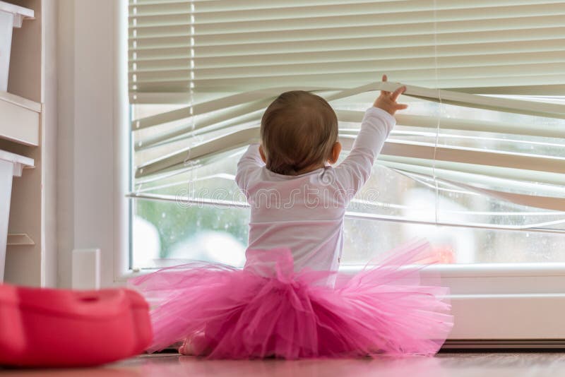 Cute little baby girl wearing a pink tutu lifting the blinds on the window so she can peer outside at the garden in a low angle rear view. Cute little baby girl wearing a pink tutu lifting the blinds on the window so she can peer outside at the garden in a low angle rear view.