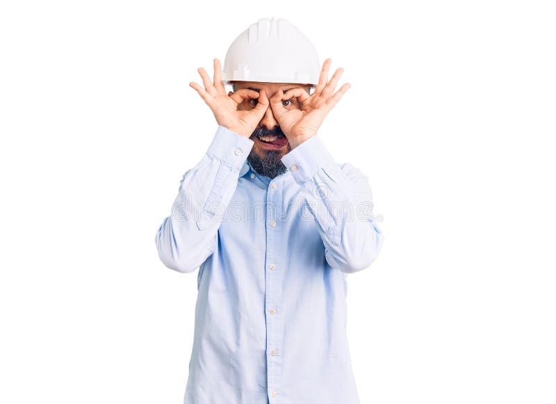 穿着建筑师硬帽的年轻帅哥做着像双筒望远镜一样的姿势，从手指伸出舌头看着 库存照片