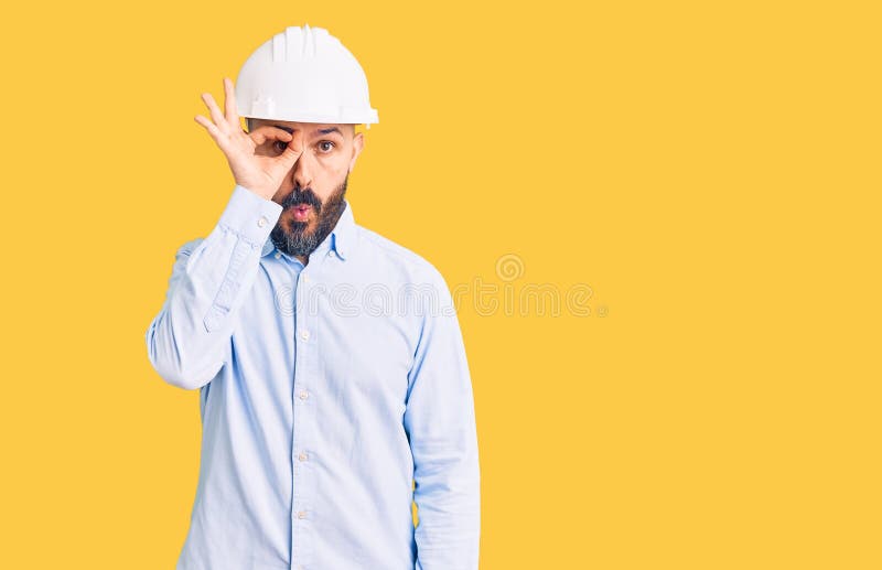穿着建筑师硬帽的年轻英俊男士，手势不错，他惊讶的面目透过手指望去 免版税库存照片