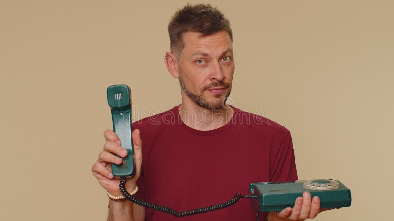 Hey you, call me back. Handsome young man in red t-shirt talking on wired vintage telephone of 80s, says hey you call me back. Call center. Adult stylish male guy on beige studio background indoor. Hey you, call me back. Handsome young man in red t-shirt talking on wired vintage telephone of 80s, says hey you call me back. Call center. Adult stylish male guy on beige studio background indoor