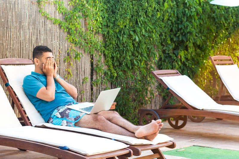 Portrait of tired handsome bearded young adult student man in blue t-shirt and shorts lying on daybed with laptop on poolside and education online, covering eyes with hand. Lifestyle, summer vacation. Portrait of tired handsome bearded young adult student man in blue t-shirt and shorts lying on daybed with laptop on poolside and education online, covering eyes with hand. Lifestyle, summer vacation